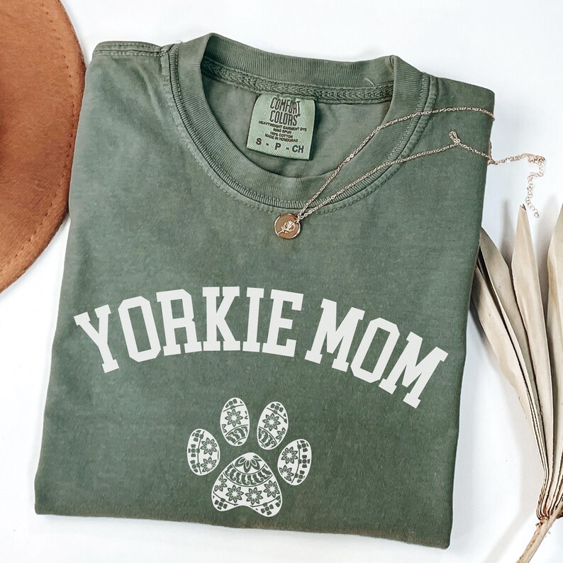 May include: An olive green t-shirt with the text "Yorkie Mom" in white block letters and a paw print design. A gold-colored necklace with a pendant rests on the shirt. The shirt is a size small.