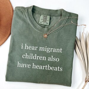 Pu&ograve; includere: Una maglietta Comfort Colors verde oliva con la scritta bianca "i hear migrant children also have heartbeats". Una collana d'oro con un ciondolo &egrave; appoggiata sulla maglietta. Un cappello marrone e una pianta secca sono sullo sfondo.