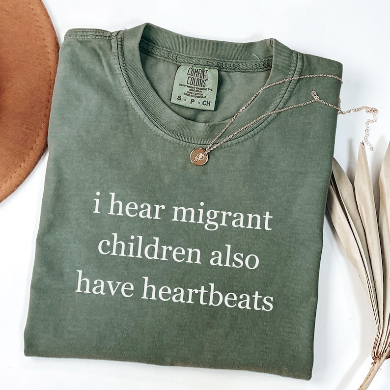 Pu&ograve; includere: Una maglietta Comfort Colors verde oliva con la scritta bianca "i hear migrant children also have heartbeats". Una collana d'oro con un ciondolo &egrave; appoggiata sulla maglietta. Un cappello marrone e una pianta secca sono sullo sfondo.