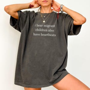Pu&ograve; includere: T-shirt grigio scuro oversize con la scritta bianca "i hear migrant children also have heartbeats". La maglietta ha maniche corte e una vestibilit&agrave; ampia, adatta per un look casual.
