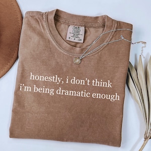 May include: A brown Comfort Colors t-shirt with the text "honestly, I don't think I'm being dramatic enough" in white. A silver necklace with a pendant is draped across the shirt. A brown hat is visible in the upper left corner.