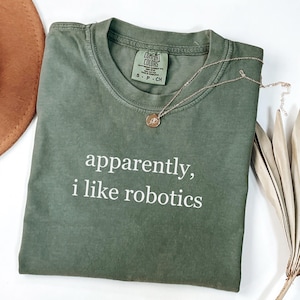 May include: An olive green short-sleeve t-shirt with a crew neckline. The shirt features the white text "apparently, i like robotics" printed on the front. A gold-toned chain necklace with a small circular pendant rests on the collar area.