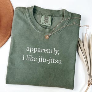May include: An olive green t-shirt with the text "apparently, i like jiu-jitsu" in white. A gold necklace with a pendant rests on the shirt. The shirt is folded and placed on a white surface.