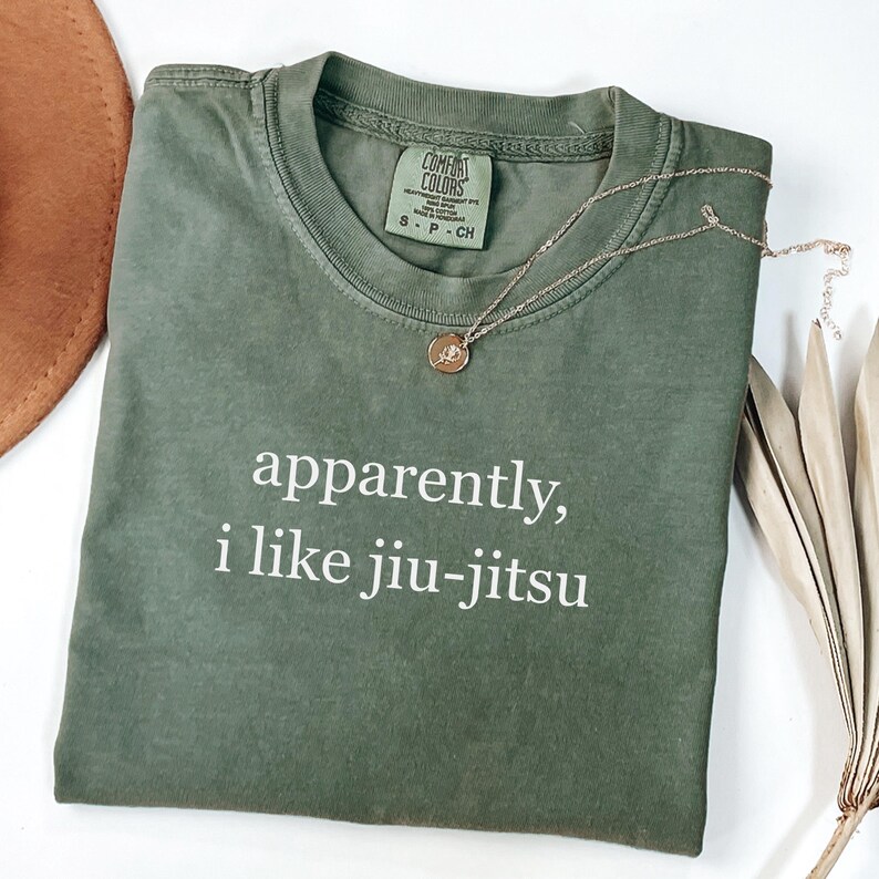 May include: An olive green t-shirt with the text "apparently, i like jiu-jitsu" in white. A gold necklace with a pendant rests on the shirt. The shirt is folded and placed on a white surface.