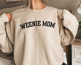 May include: A sand-colored crewneck sweatshirt featuring the text "WEENIE MOM" printed in a bold, black, arched serif font across the chest. The garment has long sleeves with ribbed cuffs and a ribbed neckline, providing a relaxed fit.