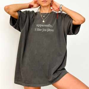 May include: Dark gray t-shirt with the text "apparently, i like jiu-jitsu" in white. The shirt has a relaxed fit and short sleeves. The image shows a person wearing the shirt.