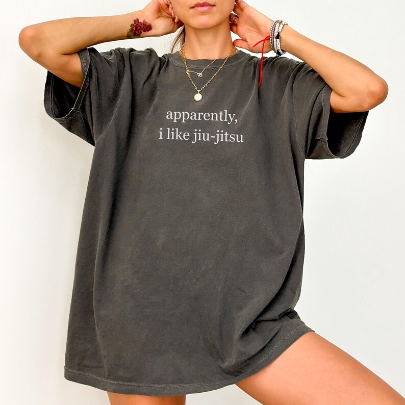 May include: Dark gray t-shirt with the text "apparently, i like jiu-jitsu" in white. The shirt has a relaxed fit and short sleeves. The image shows a person wearing the shirt.