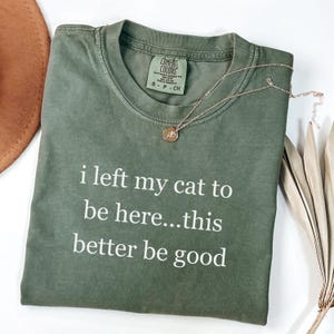 May include: An olive green t-shirt with the text "i left my cat to be here...this better be good" in white. A gold necklace with a pendant rests on the shirt. The shirt has a Comfort Colors tag.