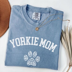 May include: A blue Comfort Colors t-shirt with the text "Yorkie Mom" in white, along with a paw print design. A gold necklace with a pendant rests on the shirt. A brown hat and dried plant stems are in the background.