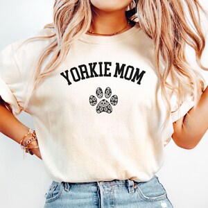 May include: Cream-colored sweatshirt with the text "YORKIE MOM" in black, arched above a paw print design. The paw print is filled with a black and white floral and star pattern. The sweatshirt is a casual style.
