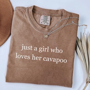 May include: A brown Comfort Colors t-shirt with the text "just a girl who loves her cavapoo" in white. A silver necklace with a small pendant is draped across the shirt. A brown hat is visible in the upper left corner.