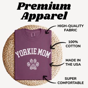 May include: A maroon t-shirt with the text "YORKIE MOM" in white, above a white paw print design. The shirt is made of 100% cotton and is labeled as high-quality fabric, made in the USA, and super comfortable. The words "Premium Apparel" are at the top.