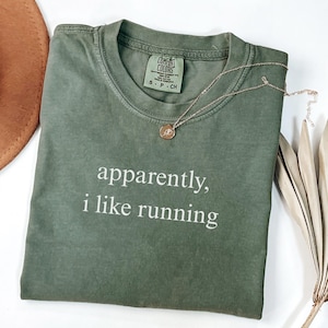 May include: An olive green t-shirt with the phrase "apparently, i like running" printed in white. A gold necklace with a pendant rests on the shirt. The shirt is folded, and a brown hat is partially visible.