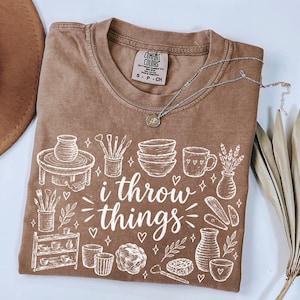 May include: A brown short-sleeve t-shirt with a white graphic print featuring pottery tools, bowls, vases, and the text "i throw things." The shirt has a Comfort Colors brand label at the neck and is paired with a silver-toned chain necklace.