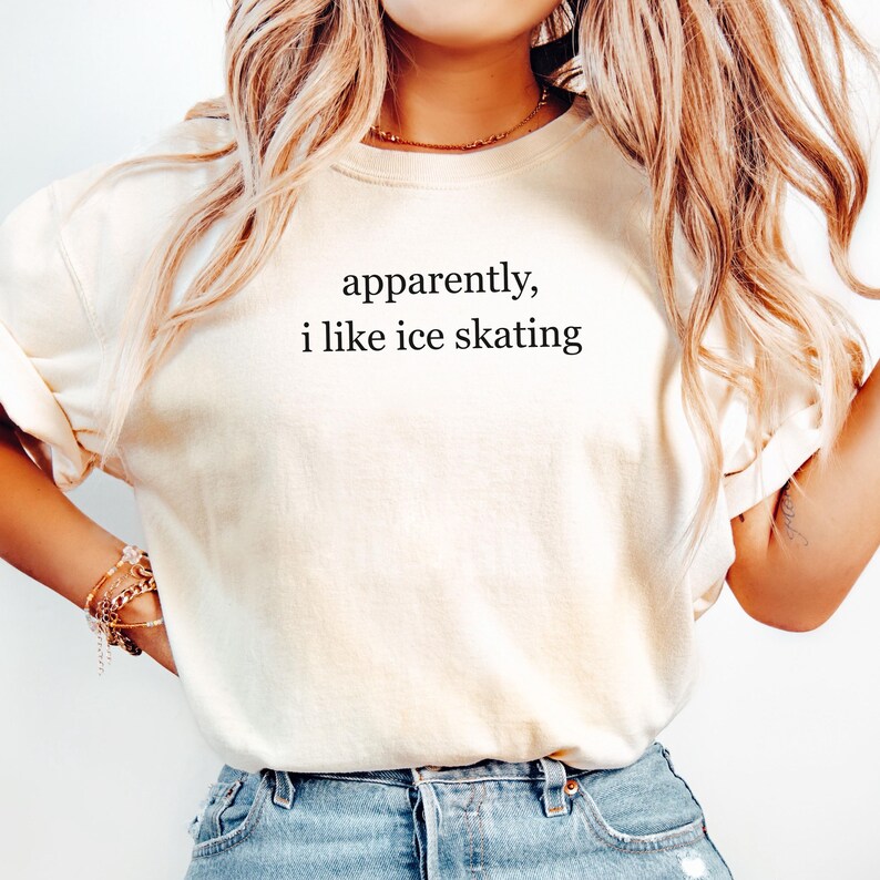 May include: Cream-colored t-shirt with the text "apparently, i like ice skating" in black. The shirt is a casual style, suitable for everyday wear. The model is wearing blue jeans.
