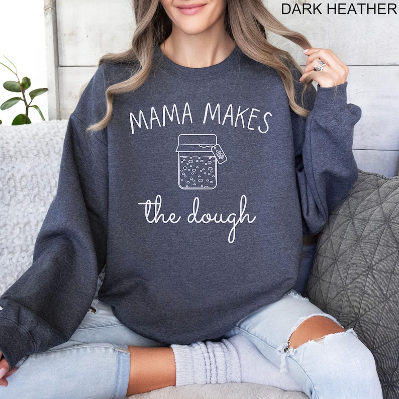 May include: A dark heather grey sweatshirt with the text "Mama Makes the Dough" in white. A white outline of a jar with a tag that says "Dough" is in the center of the text.