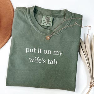 May include: An olive green t-shirt with the phrase "put it on my wife's tab" printed in white. A gold necklace with a circular pendant rests on the shirt. The shirt is folded on a white surface.