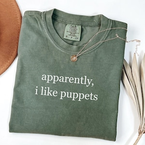 May include: An olive green t-shirt with the text "apparently, i like puppets" in white. A gold necklace with a pendant rests on the shirt. The shirt is a Comfort Colors brand, size S-P-CH.