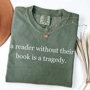 May include: An olive green t-shirt with the text "a reader without their book is a tragedy." A gold-colored necklace with a pendant rests on the shirt. The shirt has a Comfort Colors tag.