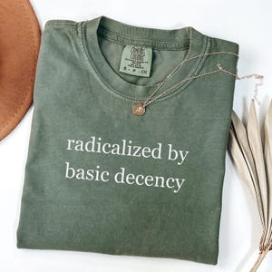 May include: An olive green t-shirt with the phrase "radicalized by basic decency" in white lettering. A gold necklace with a pendant rests on the shirt. The shirt is folded, with a brown hat and dried plant in the background.