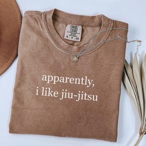 May include: A brown Comfort Colors t-shirt with the text "apparently, i like jiu-jitsu" in white. A silver necklace with a circular pendant rests on the shirt. The shirt is folded and placed on a light-colored surface.
