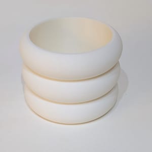 May include: A stack of three smooth, matte white circular bangles. The bracelets feature a thick, rounded, tube-like profile with a uniform, solid off-white colour throughout. The three identical pieces are stacked vertically to form a single unit.