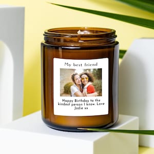 May include: A cylindrical amber glass candle jar featuring a white rectangular label. The label displays a photograph of two people embracing, with the text "My best friend" above and "Happy Birthday to the kindest person I know. Love Jodie xx" below.