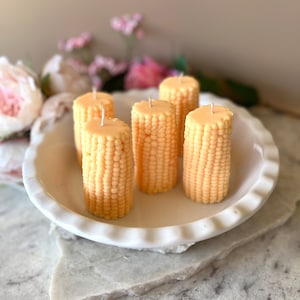May include: Six yellow corn on the cob candles in a white pie dish. The candles are arranged in a circle and have a white wick.