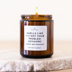 May include: A lit soy wax candle in a brown glass jar with a white label that reads Kindled Flames, SMELLS LIKE IT'S NOT YOUR PROBLEM ANYMORE! HAPPY RETIREMENT, SOY WAX CANDLE. The candle features a single flame burning at the top of the wax.