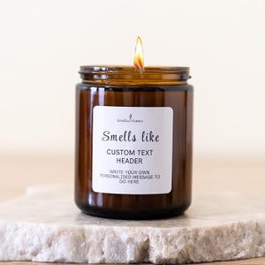 May include: An amber glass candle jar with a burning flame and a white label. The label says "Smells like", "CUSTOM TEXT HEADER", and "WRITE YOUR OWN PERSONALISED MESSAGE TO GO HERE". The candle is on a stone surface.