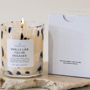 May include: A soy wax candle in a clear glass jar with black speckles, featuring a lit wick and a white label that reads: Kindled Flames, SMELLS LIKE YOU'RE ENGAGED, ENJOY THE FRESH AND INVIGORATING SCENT OF BEING OFF THE MARKET, SOY WAX CANDLE.