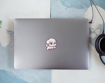 Op de afbeelding: Een zilveren laptop met een witte cartoon-hondensticker op de deksel. De laptop staat op een lichtblauw en wit oppervlak.