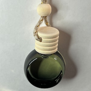 May include: A small, round, dark green glass bottle with a wooden cap and a braided beige cord. The cord has gold accents and is attached to a wooden bead. The bottle is designed to hang and is likely an air freshener or diffuser.