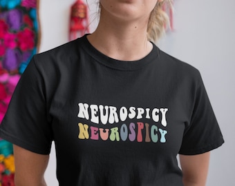 Puede incluir: Camiseta negra con la palabra "NEUROSPICY" en blanco y "NEUROSPICY" en colores pastel. La camiseta la lleva una persona. El fondo incluye tapices de pared coloridos.