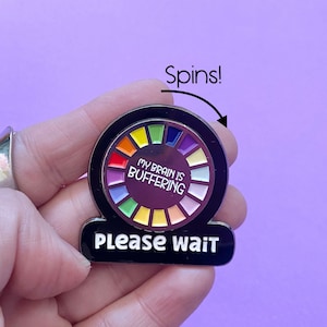 May include: A black and silver enamel pin with a spinning rainbow wheel and the text "My brain is buffering. Please wait." The wheel is spinning clockwise.