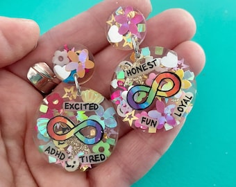 Könnte beinhalten: Ein Paar bunte Harzohrringe mit einem Regenbogen-Unendlichkeitszeichen, Glitzer und Wörtern wie "aufgeregt", "ehrlich", "loyal", "Spaß", "ADHS" und "müde".