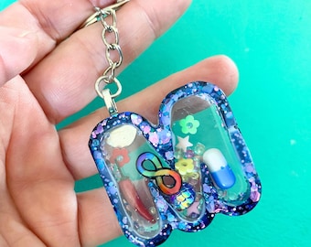 May include: A blue and purple glitter resin keychain shaped like the letter N. The keychain is filled with colorful confetti, glitter, and a rainbow infinity symbol.