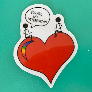 Op de afbeelding: Een sticker met twee stokpoppetjes die op een rood hart zitten. Het hart heeft een regenboog boog aan de linkerkant. Het stokpoppetje links houdt een spreekballon vast met de tekst "Jij bent mijn hyperfixatie."