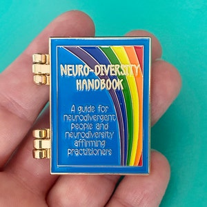 May include: A gold-colored pin shaped like a book with a blue cover and a rainbow design. The text on the cover reads "Neuro-Diversity Handbook" and "A guide for neurodivergent people and neurodiversity affirming practitioners".
