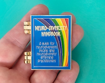 May include: A gold-colored pin shaped like a book with a blue cover and a rainbow design. The text on the cover reads "Neuro-Diversity Handbook" and "A guide for neurodivergent people and neurodiversity affirming practitioners".
