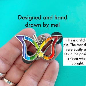 May include: A rainbow-colored butterfly mask pin with a silver star that slides up and down. The pin is designed and hand-drawn by the artist. "This is a sliding pin. The star slides very easily and sits in the position shown when upright."