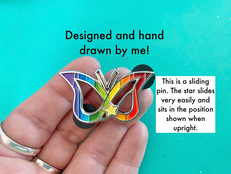 May include: A rainbow-colored butterfly mask pin with a silver star that slides up and down. The pin is designed and hand-drawn by the artist. "This is a sliding pin. The star slides very easily and sits in the position shown when upright."