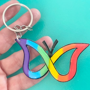 Könnte beinhalten: Ein regenbogenfarbener Schmetterlings-Schlüsselanhänger mit einem silbernen Schlüsselring. Der Schmetterling ist aus Acryl und hat einen schwarzen Umriss.