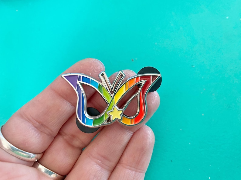 May include: A rainbow-colored butterfly pin with a yellow star in the center. The pin is made of metal and has a silver finish.