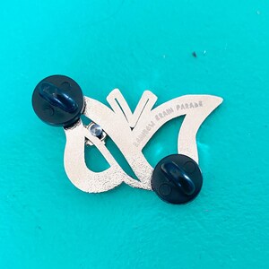 May include: Silver butterfly pin with black rubber backings. The pin features the text "Rainbow Brain Parade" on the back.
