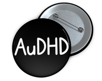 May include: A black button with a white inscription that reads "AuDHD". The button is attached to a silver pin back.