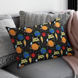 Puede incluir: Una almohada negra y blanca con un patr&oacute;n repetido de planetas, estrellas y el texto "SAFE SPACE".
