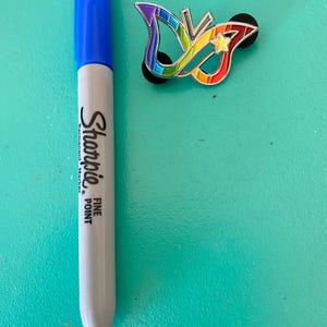 May include: A rainbow butterfly pin with a star in the center. The pin is on a turquoise surface next to a blue and gray Sharpie fine point permanent marker.