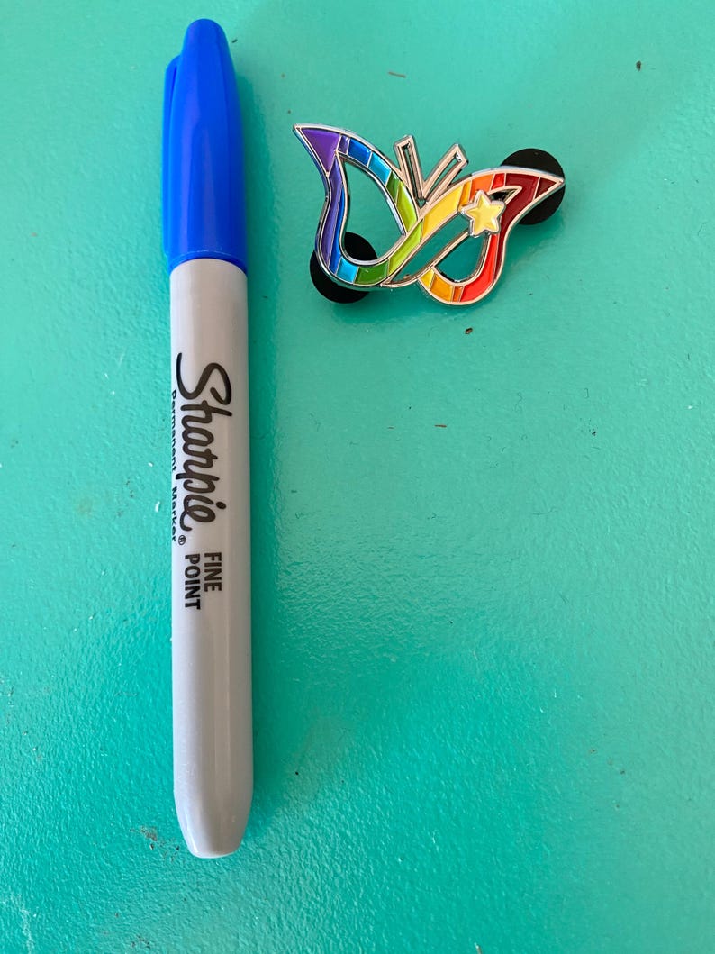 May include: A rainbow butterfly pin with a star in the center. The pin is on a turquoise surface next to a blue and gray Sharpie fine point permanent marker.