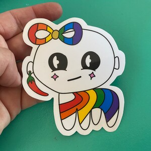 May include: A white sticker featuring a cartoon character with a rainbow bow, rainbow legs, and a red chili pepper earring. The character has large black eyes, a neutral expression, and pink star accents.